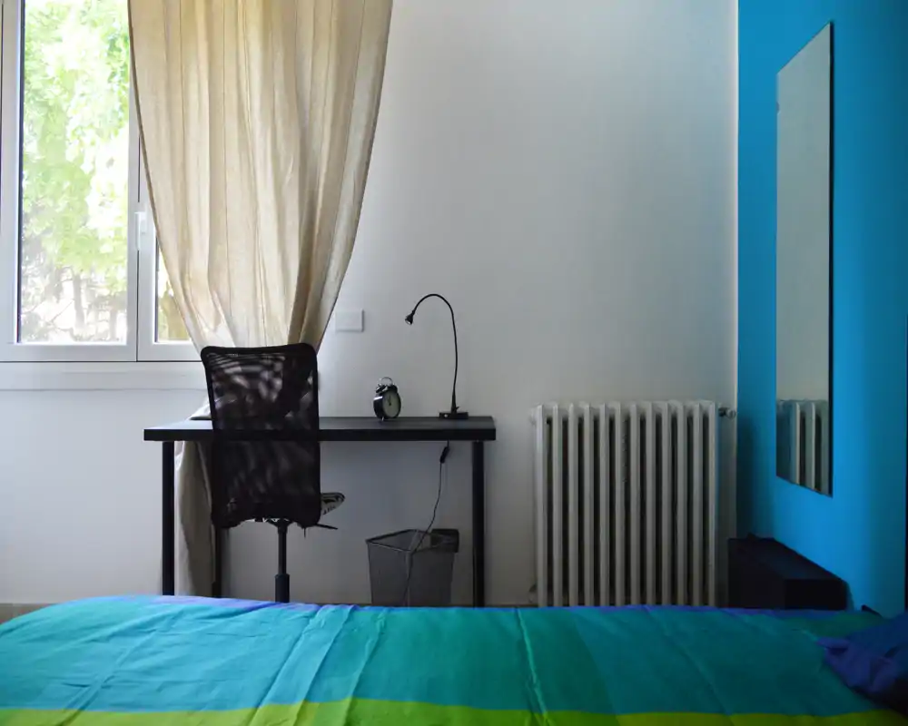 Dormitorio luminoso y sencillo con cama de colores en primer plano, un pequeño escritorio y silla junto a la ventana y un espejo de cuerpo entero en pared azul.