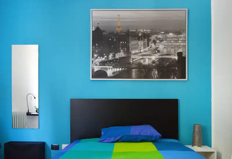 Dormitorio moderno y luminoso con colcha colorida, cabecero oscuro y una gran foto en blanco y negro de la ciudad sobre la cama. Incluye un espejo vertical y una mesita con una lámpara decorativa; diseño sencillo y atractivo para un anuncio.