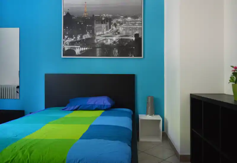 Dormitorio moderno y luminoso con edredón azul y verde, cabecero negro y mesita sencilla. Sobre la cama hay una gran fotografía en blanco y negro de la ciudad y mobiliario minimalista.