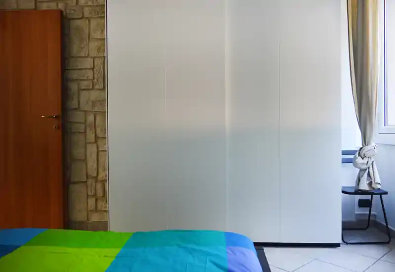 Dormitorio luminoso y sencillo con colcha colorida, gran armario blanco y ventana con cortinas anudadas que dejan entrar luz natural.