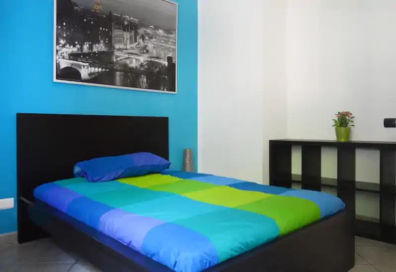 Dormitorio moderno y luminoso con cama doble colorida, cabecero negro simple y estantería minimalista — ordenado y listo para huéspedes.