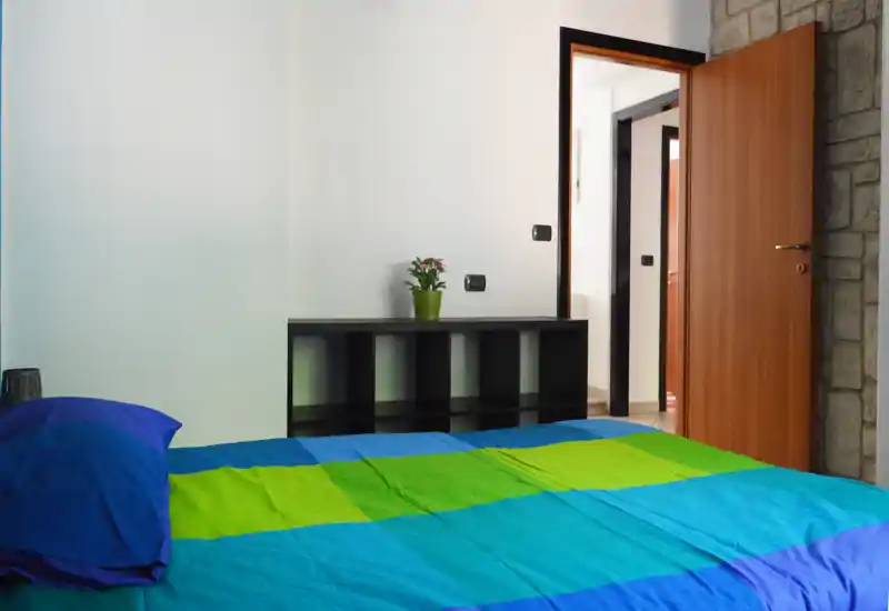 Dormitorio luminoso y moderno con una cama doble colorida en primer plano, una estantería baja con una planta en maceta y una puerta de madera abierta hacia el pasillo.