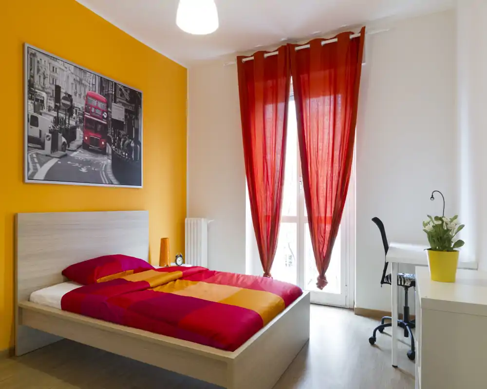 Chambre moderne et lumineuse avec grand lit aux couettes rouges et orange, mur d'accent coloré et bureau près de la fenêtre — idéale pour les locations courte durée.