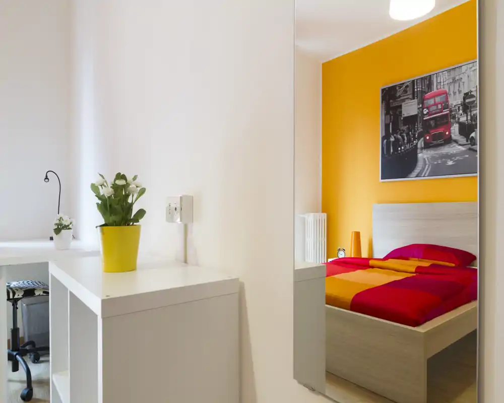 Chambre moderne et lumineuse principalement visible dans un miroir mural, avec une couette rouge et orange et un cadre de lit en bois clair ; décor épuré et mur d'accent coloré.