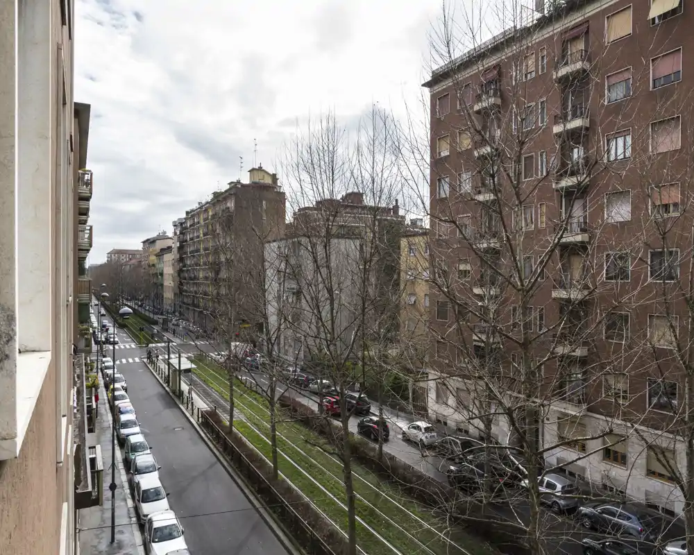 Vue sur la rue et les voies de tram depuis une fenêtre ou un balcon à l'étage; immeubles, voitures stationnées et arbres dépouillés bordent l'avenue sous un ciel couvert.