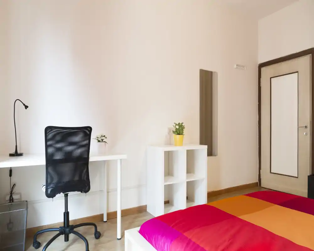 Chambre lumineuse et moderne avec une couette colorée, un bureau et une chaise, ainsi que des étagères simples et des plantes — idéale pour une personne ou un étudiant.