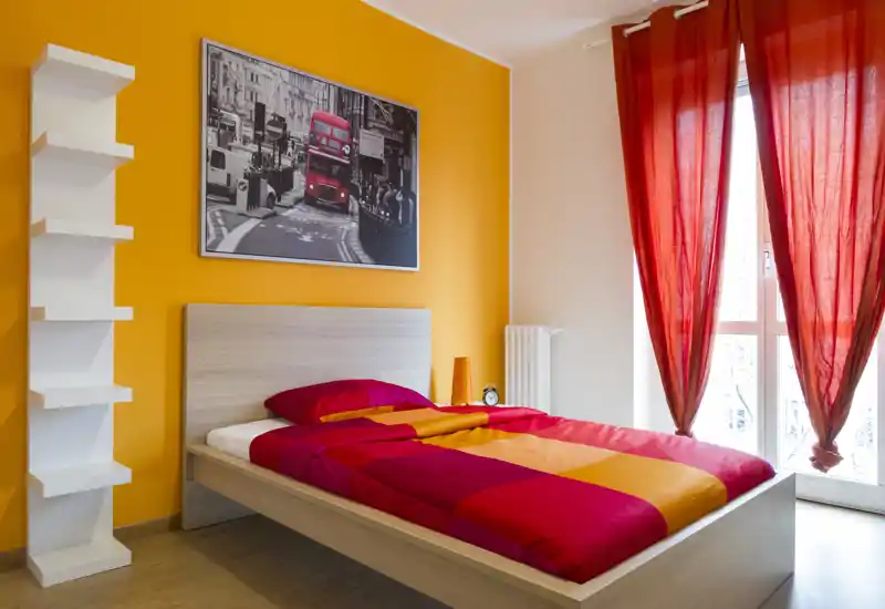 Dormitorio moderno y luminoso con cama doble frente a una pared naranja y una gran ventana con cortinas rojas. Ropa de cama cálida, lámpara de noche y un cuadro decorativo, ideal para fotos de alquiler.