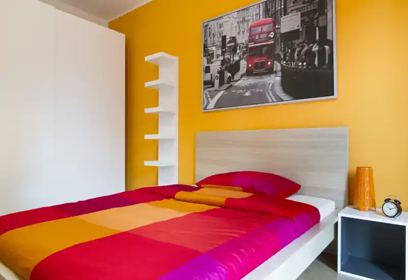 Dormitorio luminoso y colorido con una cama grande vestida con ropa de cama roja y naranja, cabecero de madera, mesita y cuadro decorativo.