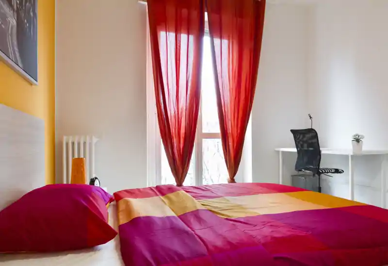 Dormitorio luminoso y colorido con cama doble vestida en tonos rojos y naranjas, ventana de suelo a techo con cortinas rojas y un pequeño escritorio con silla.