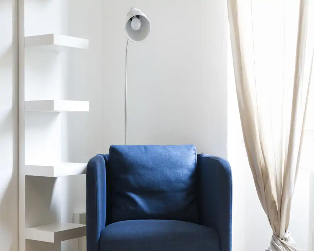 Rincón de salón luminoso y minimalista con un sillón tapizado azul intenso, lámpara de pie y suave luz natural desde la ventana — un acogedor espacio de lectura.