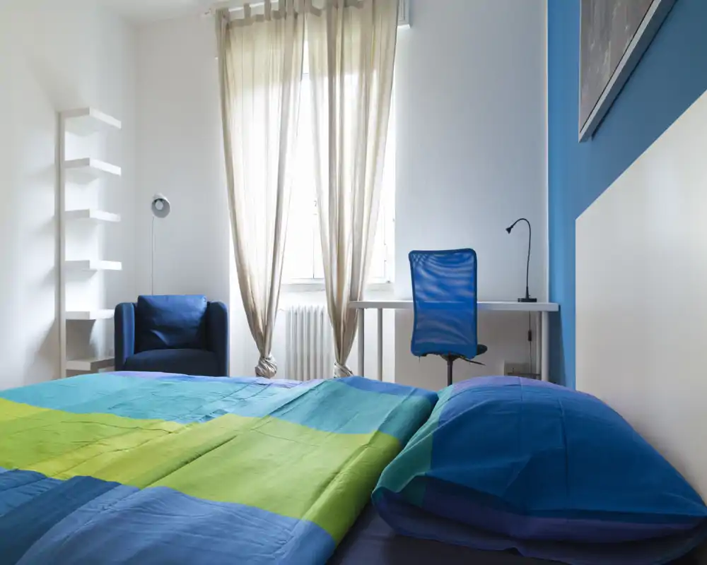 Dormitorio luminoso y moderno con ropa de cama colorida, escritorio y silla junto a la ventana y un sillón cómodo — ideal para descansar o trabajar.