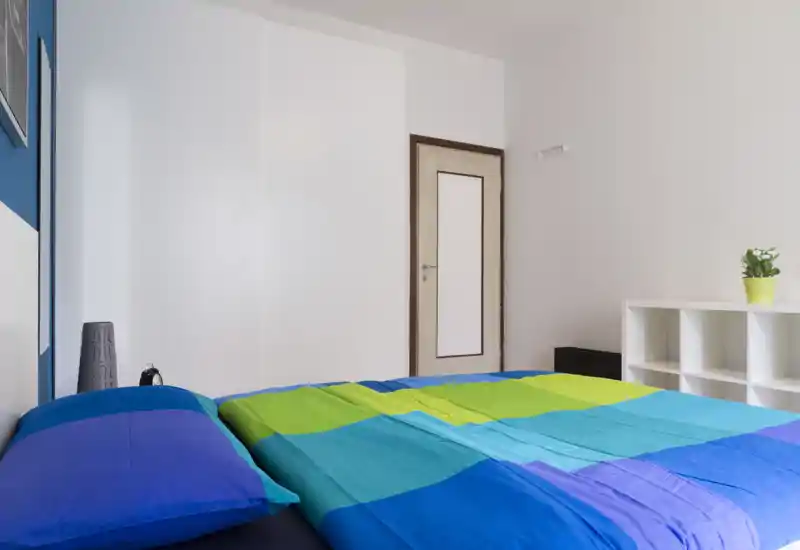 Dormitorio moderno y luminoso con una amplia cama colorida que domina la imagen, paredes blancas y estantería minimalista; perfecto para destacar la zona de descanso.