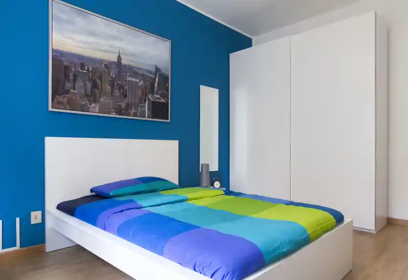 Dormitorio moderno con cama blanca baja y ropa de cama azul-verde, armario grande y pared azul de acento; luminoso, ordenado y bien compuesto para anuncios.