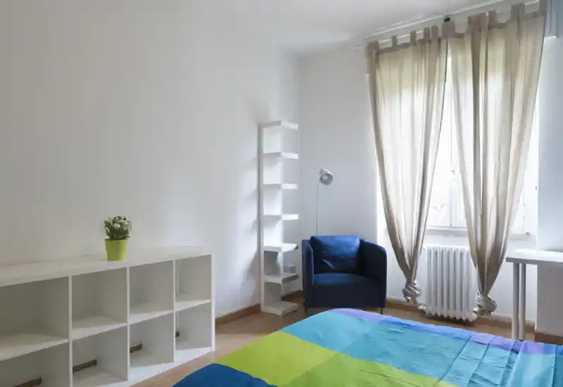 Dormitorio luminoso y sencillo con una colcha colorida, un sillón azul junto a la ventana y estanterías minimalistas. La luz natural y las líneas limpias crean una sensación fresca y aireada, perfecta para alquileres de corta duración.