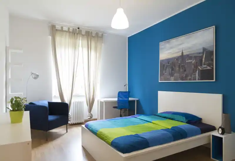 Dormitorio moderno y luminoso con cama blanca, ropa de cama colorida y pared azul intensa. La habitación incluye escritorio, sillón y una gran ventana con cortinas, creando un espacio cómodo y bien iluminado para alquileres.