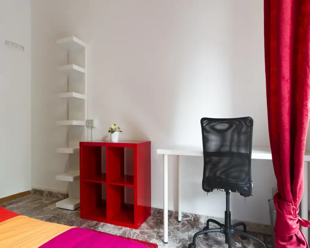 Rincón de dormitorio luminoso y ordenado con escritorio y silla de oficina de malla, ropa de cama colorida y una pequeña estantería roja; ideal para una persona o estudiante.