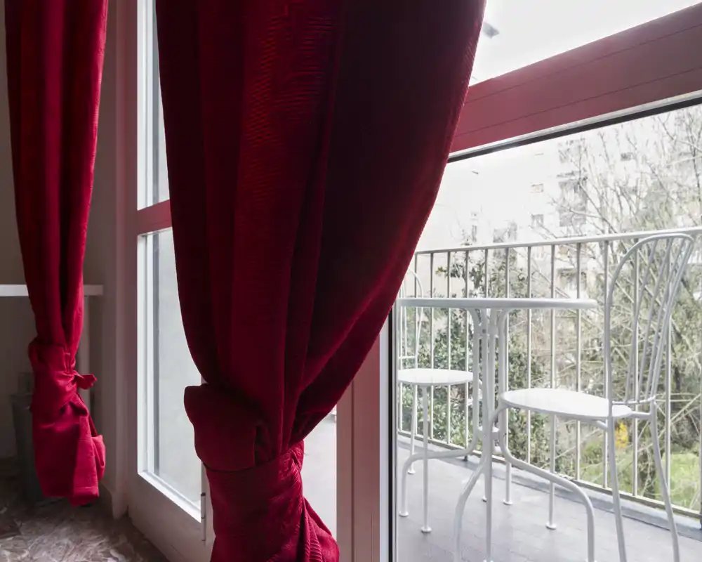 Pequeño balcón con mesa tipo bistró de metal blanco y dos sillas, visto a través de puertas correderas enmarcadas por cortinas rojas — ideal para un café matutino.