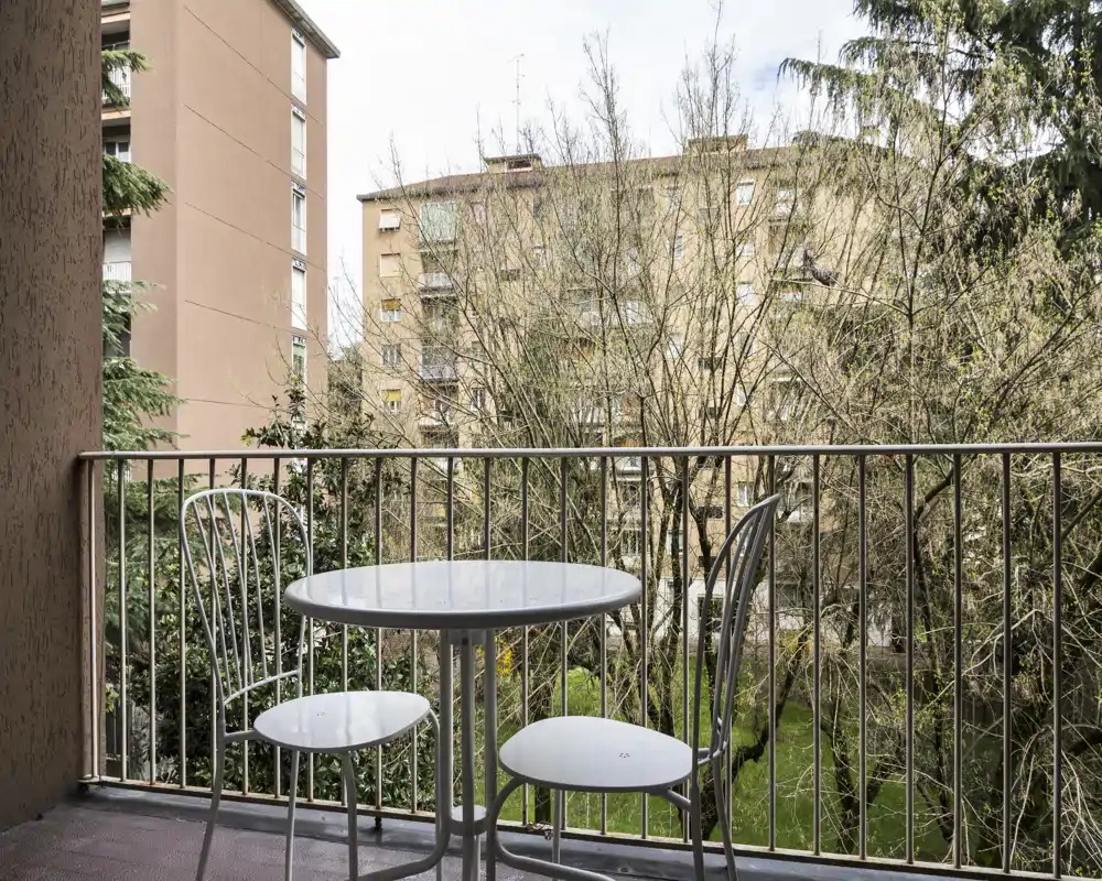 Pequeño balcón amueblado con mesa redonda y dos sillas de metal con vistas a árboles y edificios cercanos; un lugar agradable para el desayuno.
