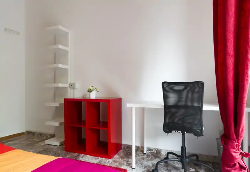 Rincón de dormitorio luminoso y ordenado con escritorio y silla de oficina de malla, ropa de cama colorida y una pequeña estantería roja; ideal para una persona o estudiante.