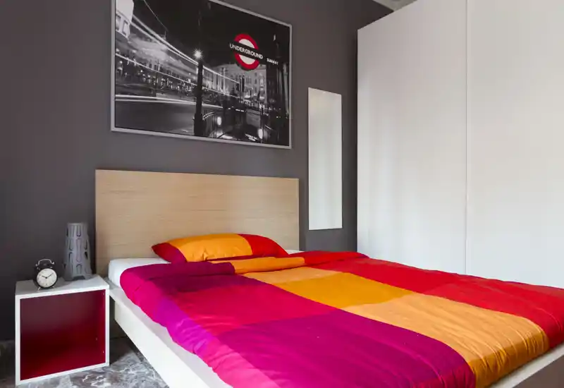 Dormitorio moderno y luminoso con cama doble grande con ropa de cama naranja y fucsia, cabecero de madera, mesita con despertador y decoración minimalista.