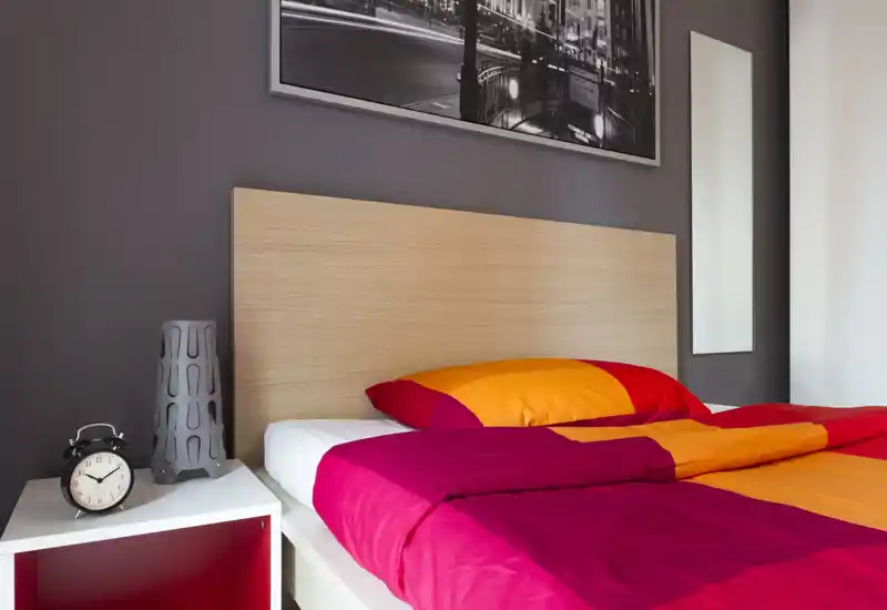 Dormitorio moderno y luminoso con cabecero de madera y ropa de cama roja y naranja; mesita con reloj y lámpara decorativa que aportan un ambiente ordenado y acogedor.