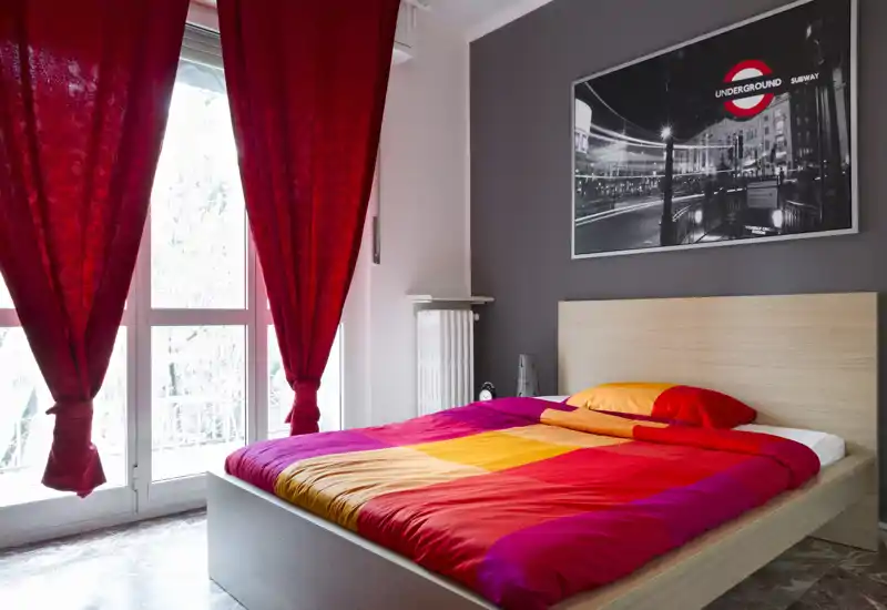 Dormitorio moderno y luminoso con una cama grande vestida con una colcha colorida, una gran ventana con cortinas rojas y un cuadro contemporáneo. La habitación resulta acogedora y bien iluminada, perfecta para un anuncio de alquiler.