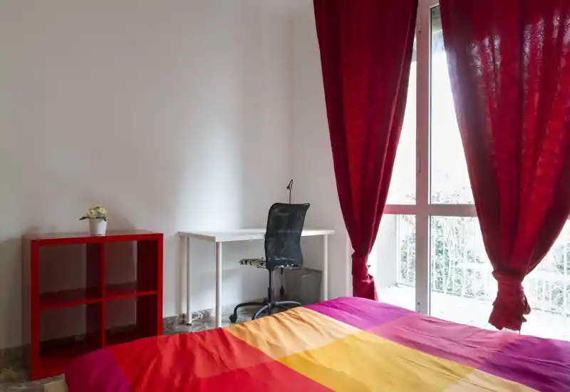 Dormitorio luminoso y colorido con una gran ventana, cortinas rojas y una cama bien hecha con ropa de cama en tonos cálidos. Un pequeño espacio de trabajo con escritorio y silla está junto a la pared y una estantería roja aporta almacenamiento y color.