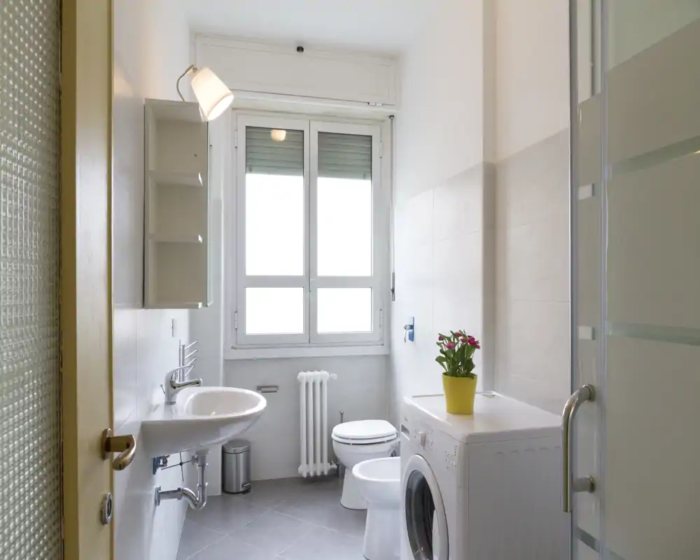 Baño moderno y luminoso con lavabo, inodoro, bidé y lavadora bajo una gran ventana; azulejos neutros y una planta aportan frescura.