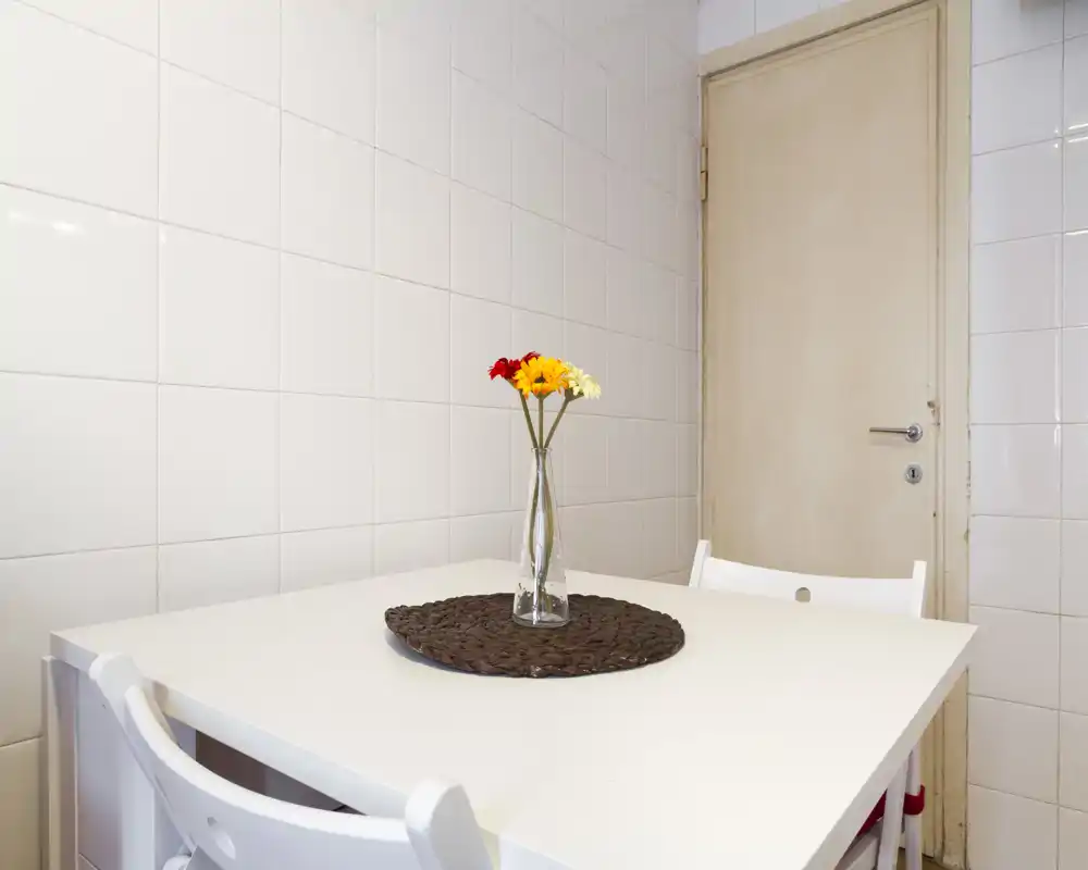 Pequeña zona de comedor luminosa con mesa blanca, dos sillas y un jarrón de cristal con flores sobre un salvamantel trenzado.