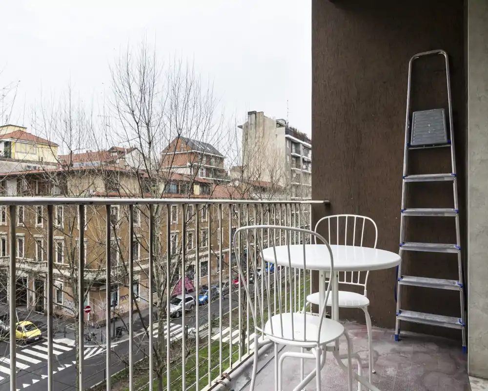 Balcón urbano con una pequeña mesa redonda y dos sillas de metal, con vistas a la calle y los edificios vecinos — un espacio exterior compacto para el café de la mañana.