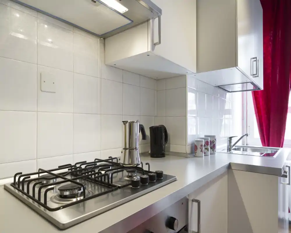 Cocina moderna y compacta con placa de gas, pequeños electrodomésticos de acero y ventana luminosa con cortina roja — práctica y bien cuidada.