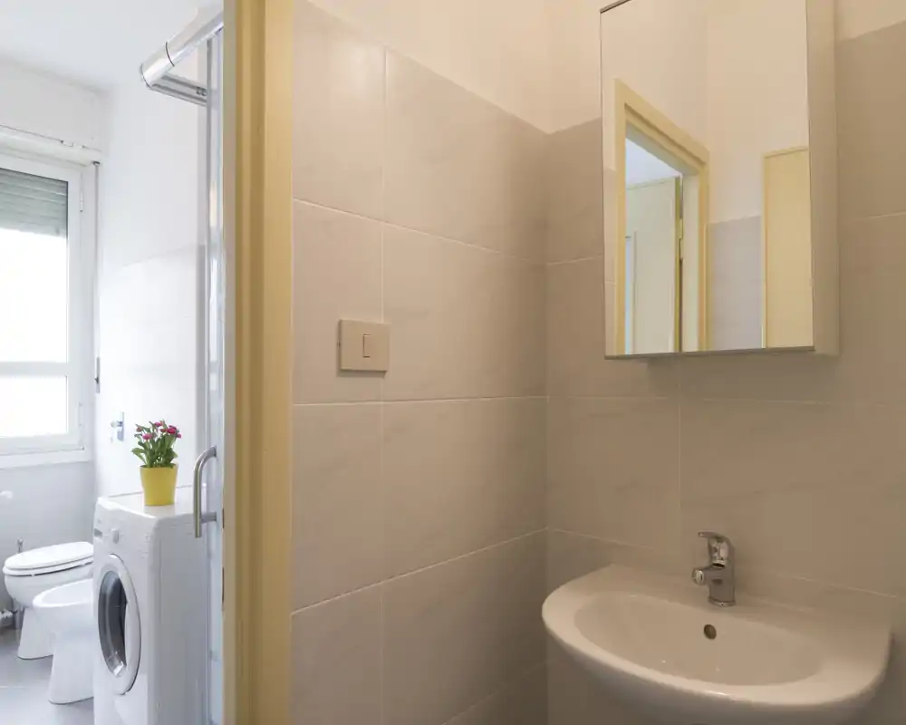 Pequeño baño moderno con lavabo de pared, armario con espejo y WC y lavadora visibles en el área contigua; azulejos neutros y luz natural suave.