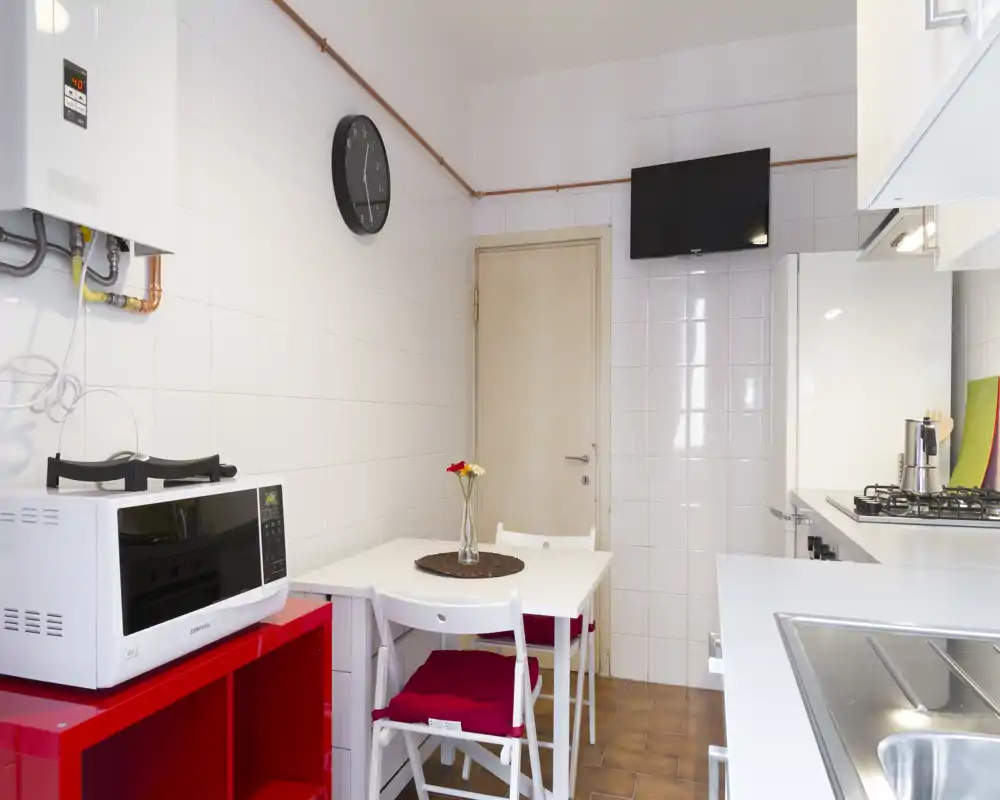 Cocina compacta amueblada con mesa pequeña, cocina de gas, microondas y fregadero. Espacio luminoso y ordenado con detalles de color y asiento para dos.