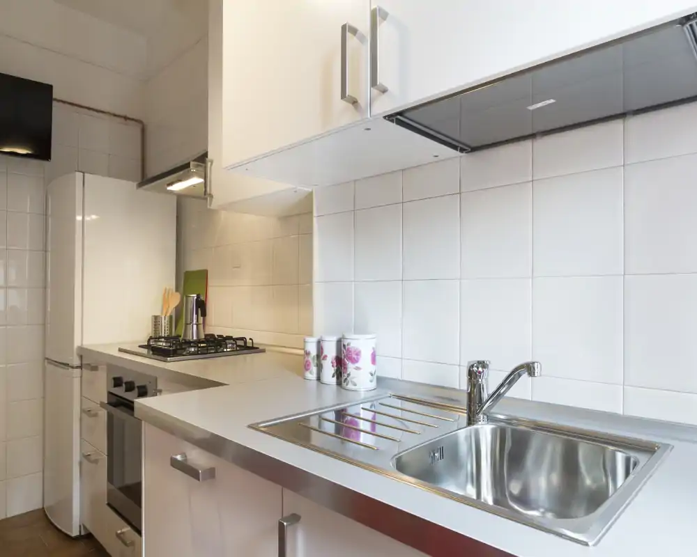 Cocina compacta y moderna con fregadero de acero inoxidable, placa de gas y muebles blancos — luminosa y ordenada para cocinar.