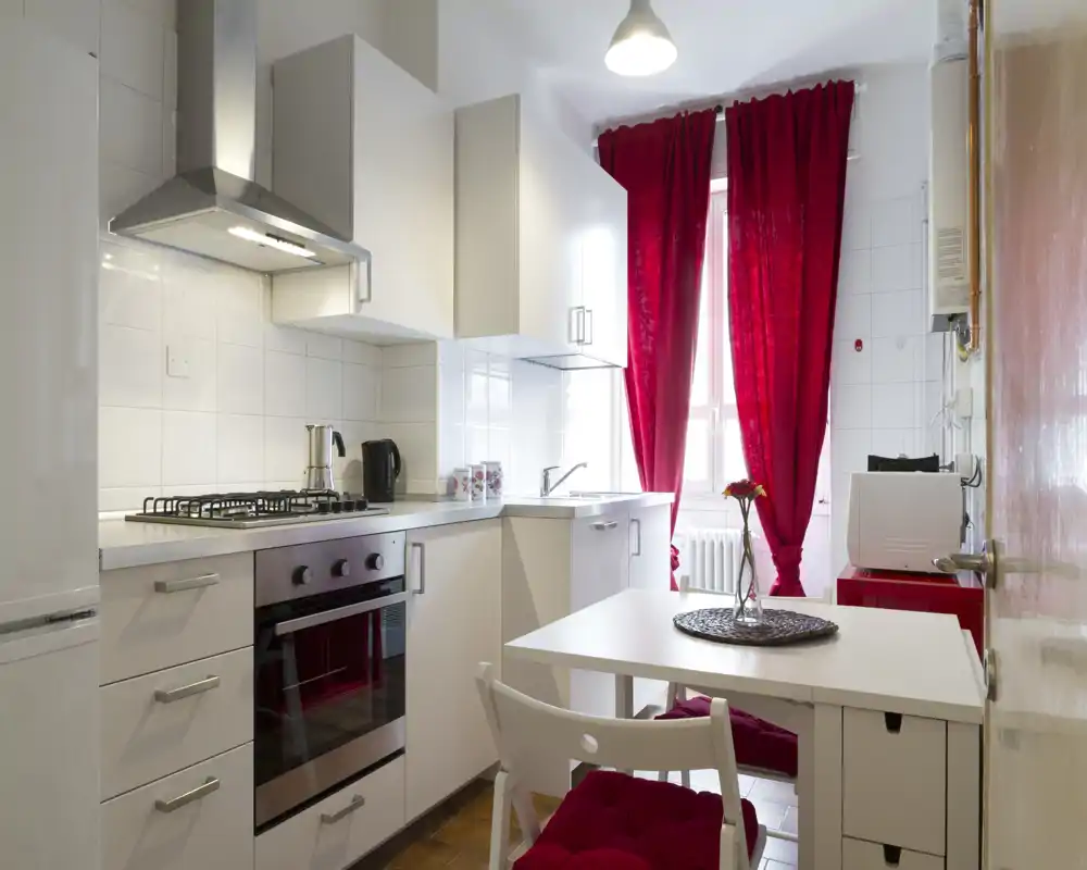 Cocina compacta y luminosa con armarios blancos modernos, cocina de gas, mesa pequeña y cortinas rojas intensas que crean un ambiente acogedor y bien iluminado.