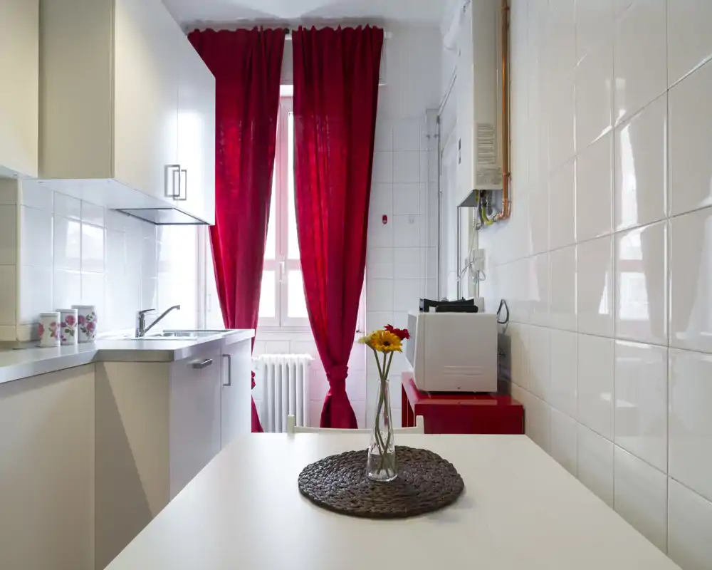 Cocina compacta y luminosa con muebles blancos, fregadero, superficie de trabajo y pequeño comedor; cortinas rojas y un jarrón aportan un acento cálido y elegante.