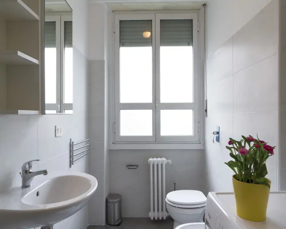 Baño moderno y luminoso con lavabo, inodoro, radiador y lavadora; azulejos blancos y una amplia ventana esmerilada que aporta mucha luz natural.