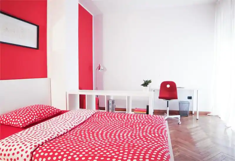 Camera da letto moderna e luminosa con letto matrimoniale completamente visibile con biancheria rossa a pois, una scrivania bianca con sedia vicino alla finestra e pavimento in parquet a spina di pesce.