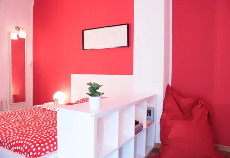 Camera da letto moderna e luminosa con pareti rosse vivaci, testiera bianca e copriletto a pois. Una libreria divisoria, lampada, pianta e un pouf rosso creano un ambiente accogliente e di stile, perfetto per annunci.