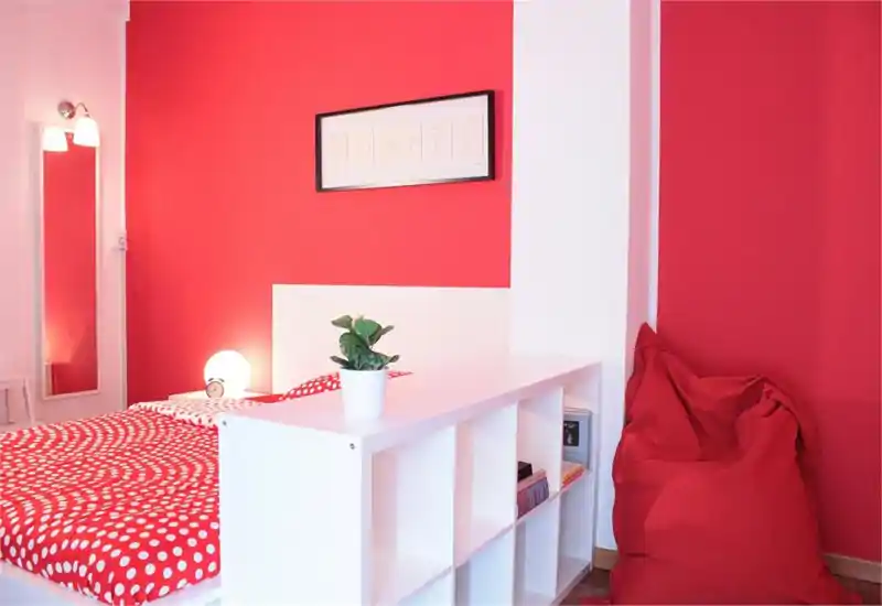 Camera da letto moderna e luminosa con struttura letto bianca, biancheria rossa a pois e un comodo puff; stile minimalista pulito e ottima illuminazione.