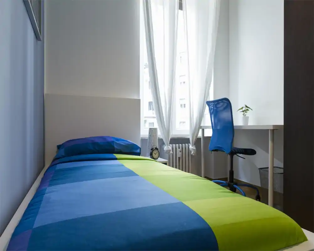 Habitación individual luminosa y moderna con colcha colorida, cabecero, mesita con despertador y un puesto de trabajo junto a la ventana. La luz natural y el estilo minimalista la hacen ideal para estudiantes o huéspedes individuales.