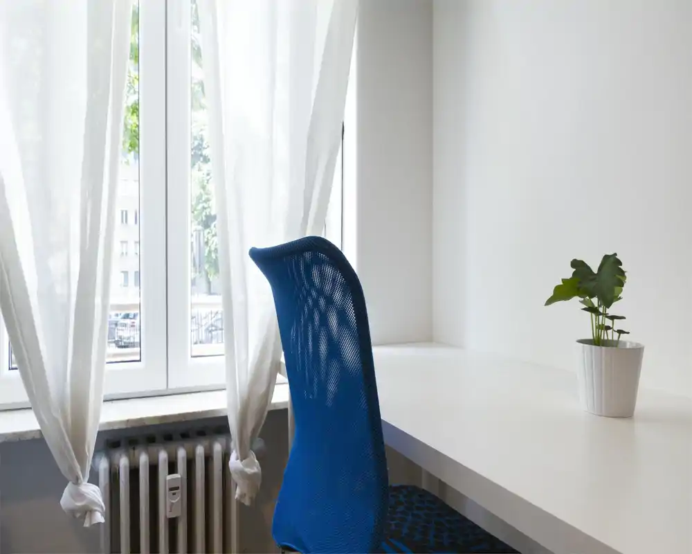 Rincón luminoso y minimalista junto a una gran ventana con cortinas traslúcidas, silla azul de malla y escritorio blanco con una pequeña maceta: ideal como espacio de trabajo o lectura.