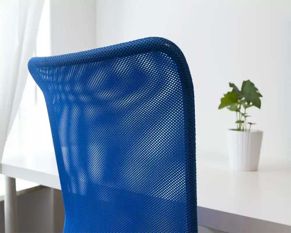 Plano detalle del respaldo de malla azul de una silla de oficina frente a un escritorio blanco con una pequeña planta en maceta; mínimo contexto de la habitación.