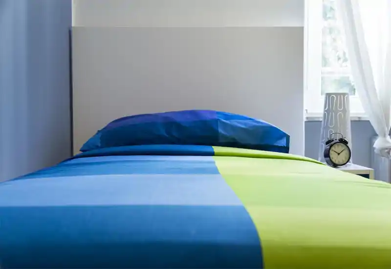 Dormitorio luminoso y moderno con cama bien hecha en ropa azul y verde, cabecero minimalista y mesita con reloj despertador junto a la ventana.