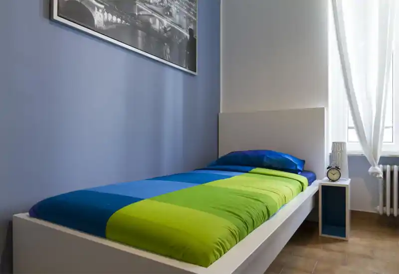 Dormitorio individual moderno y luminoso con ropa de cama azul y verde llamativa, cabecero blanco sencillo y mesita junto a una ventana que aporta luz natural.