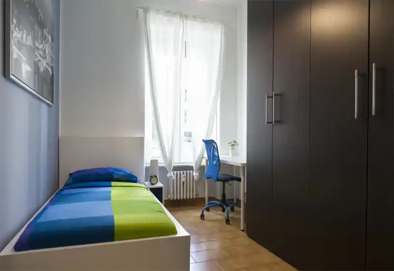 Dormitorio individual luminoso y ordenado con la cama completamente visible, escritorio junto a la ventana y un gran armario oscuro — moderno y bien iluminado.