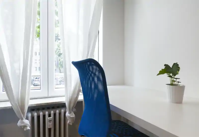 Rincón luminoso y minimalista junto a una gran ventana con cortinas traslúcidas, silla azul de malla y escritorio blanco con una pequeña maceta: ideal como espacio de trabajo o lectura.