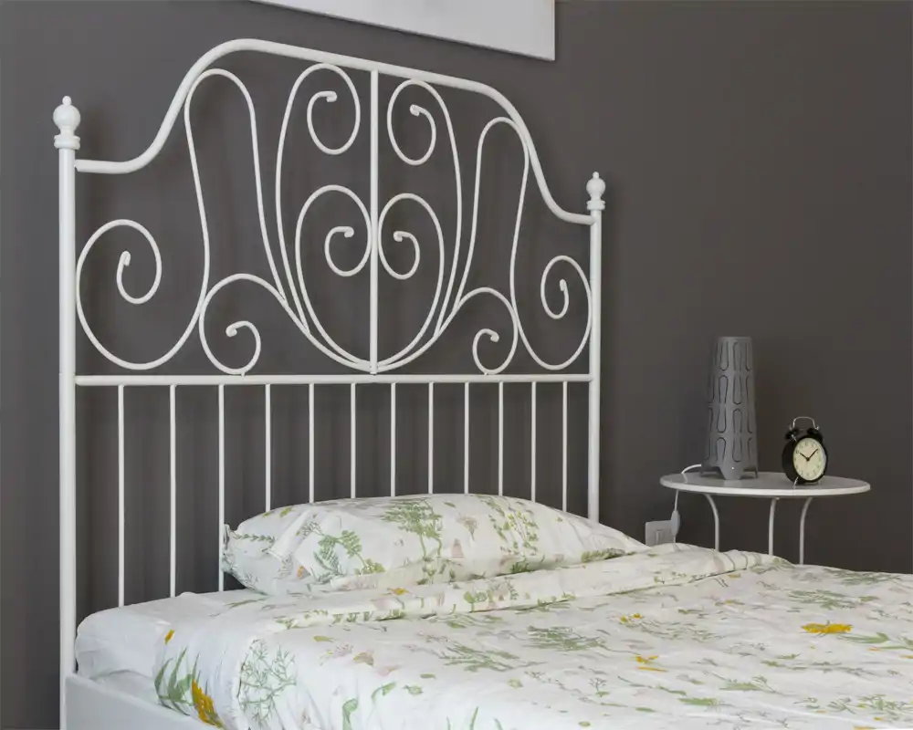 Dormitorio luminoso y ordenado con cabecero de metal blanco decorativo, ropa de cama floral y mesita con lámpara y despertador — perfecto para un anuncio acogedor.