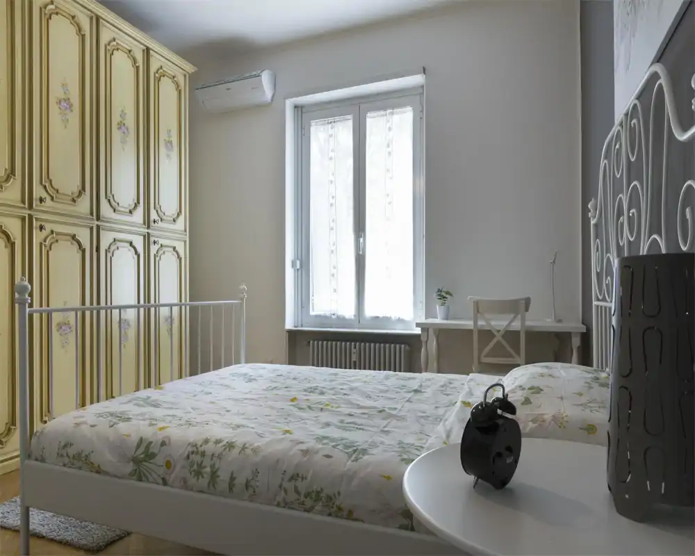 Dormitorio luminoso y acogedor con cama de metal blanca, edredón floral y un gran armario de estilo vintage. La luz natural entra por la ventana central; hay un pequeño escritorio y una silla a un lado.
