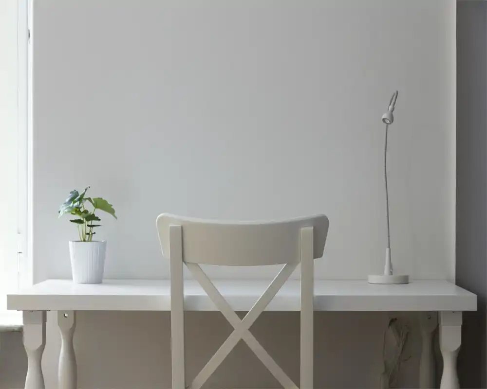 Zona de escritorio minimalista con silla blanca, lámpara sencilla y una pequeña planta en maceta junto a una ventana luminosa, que ofrece un espacio de trabajo sereno.